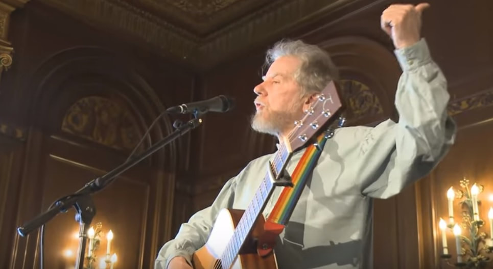 Irish singer Tommy Sands telling a story on stage at Monmouth University while holding his guitar